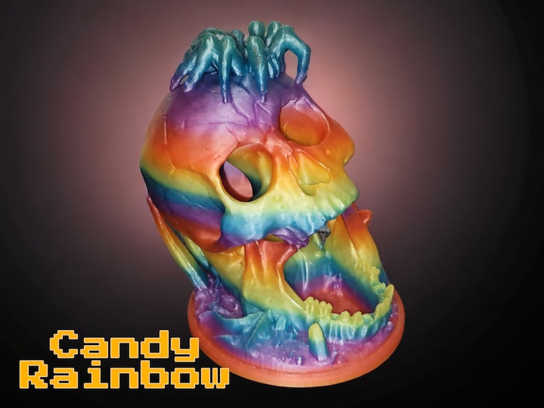May include: A colorful skull sculpture with a rainbow gradient of colors, including red, orange, yellow, green, blue, and purple. The skull has an open mouth and is topped with several small, blue figures. The text "Candy Rainbow" is displayed in a pixelated font.