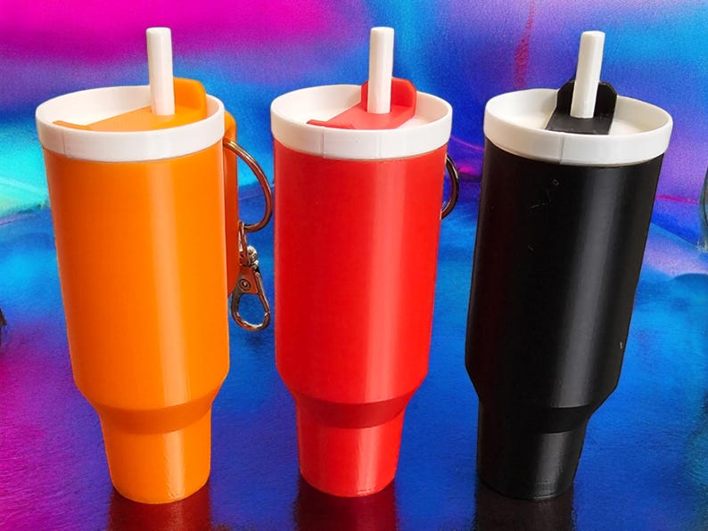 May include: Three reusable beverage containers with attached keychains. The containers are orange, red, and black. Each container has a white lid with a built-in straw.