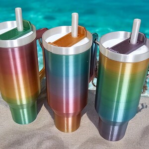 May include: Three stainless steel travel mugs with rainbow ombre finishes. Each mug has a lid with a built-in straw and a handle.