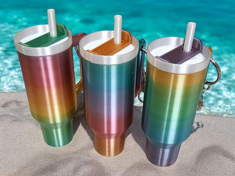 May include: Three stainless steel travel mugs with rainbow ombre finishes. Each mug has a lid with a built-in straw and a handle.