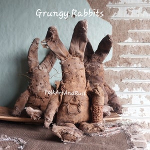 Peut inclure: Trois lapins en tissu marron avec de longues oreilles et des détails cousus. Les lapins ont un aspect vieilli et vintage. Le texte "Grungy Rabbits" et "FolkArtAndRust" est visible sur l'image, suggérant un article artisanal.