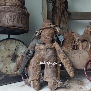 May include: A handcrafted scarecrow doll with a hat and distressed burlap clothing. The doll has a rustic, aged look. A burlap decoration with the word "HARVEST" is visible. The text "Primitive Scarecrow Gingerbread Man & Crow" is also visible.