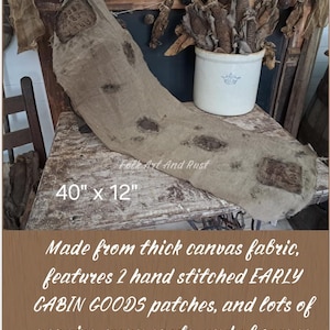 May include: A tan canvas table runner measuring 40 inches by 12 inches. The runner features two hand-stitched patches labeled EARLY CABIN GOODS, along with multiple visible repairs, worn spots, and a distressed, textured appearance throughout.