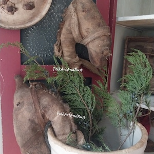 May include: A rustic, white ceramic pot filled with green branches and blue berries. Two burlap horse head decorations are placed in front of the pot. A wooden plate and other decorative items are visible in the background. The image is taken in natural light.
