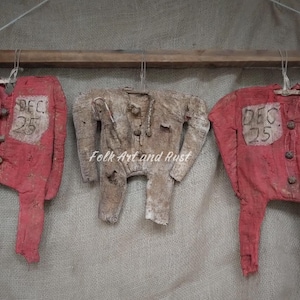 May include: Three handmade, rustic-style Christmas ornaments shaped like vintage children's pajamas. Two are red with "DEC 25" on a patch, and one is beige. They hang from a wooden rod.