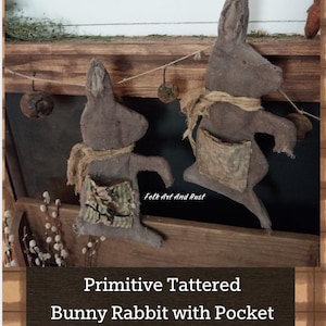 May include: Two primitive tattered bunny rabbit figures with pockets. The bunnies are made of a brown fabric and have a rustic, aged appearance. Each bunny has a burlap scarf and a pocket made of patterned fabric. The text on the image says "Primitive Tattered Bunny Rabbit with Pocket PATTERN about 36cm x 18cm".