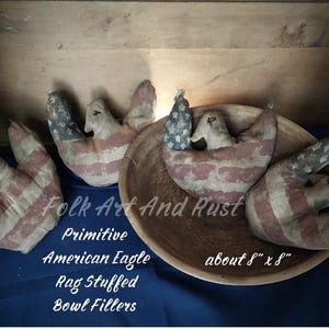 May include: Primitive rag stuffed American eagle bowl fillers, approximately 8" x 8", with a distressed American flag design. The eagles are in a wooden bowl, with the text "Folk Art And Rust" visible.