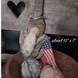 May include: A handcrafted, primitive Uncle Sam doll, approximately 12 inches by 7 inches, with a fabric hat, beard, and an American flag. The doll is seated in a wooden bowl, evoking a rustic, folk art aesthetic.