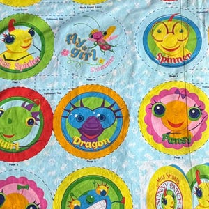 May include: A light blue fabric with colorful circular designs featuring cartoon characters. Each circle has a different character with a name like "Miss Spider" or "Dragon". The text "Fly Girl Shimmer" is also present.