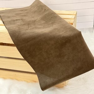 May include: A folded piece of brown suede fabric. The fabric is a solid color and has a soft, velvety texture. The suede is draped over a wooden crate, with a white, fluffy surface visible in the background.