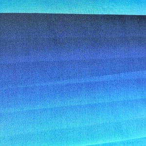 Op de afbeelding: Een stofstaal met een horizontaal blauw verloop dat overgaat van donker marineblauw naar licht cyaan. De witte zelfkant aan de linkerkant toont de zwarte gedrukte tekst NANCY CROW for JOHN KALDOR, wat de ontwerper en fabrikant aangeeft.