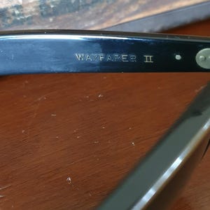 May include: Close-up of black Wayfarer II sunglasses. The text "WAYFARER II" is visible on the arm of the sunglasses. The sunglasses are on a wooden surface. The sunglasses are a classic style.