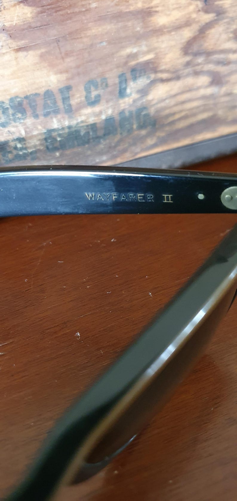 May include: Close-up of black Wayfarer II sunglasses. The text "WAYFARER II" is visible on the arm of the sunglasses. The sunglasses are on a wooden surface. The sunglasses are a classic style.