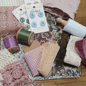 May include: A collection of sewing supplies including light blue and green buttons on cards labelled Boutique, spools of purple and green thread, various lace trims in pink and cream, a dark brown tassel, and fabric scraps with floral and plaid patterns.