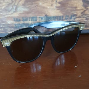 May include: Black sunglasses with a gold-colored trim on the upper frame. The lenses are a dark brown color. The sunglasses have a classic, retro design.