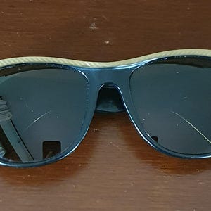 May include: Black sunglasses with dark lenses and a black frame. The top of the frame has a tan and gold striped design. The sunglasses are resting on a brown surface.