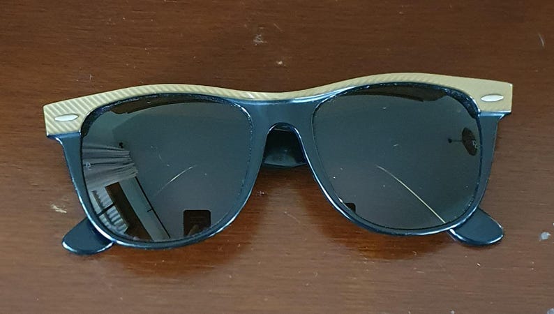 May include: Black sunglasses with dark lenses and a black frame. The top of the frame has a tan and gold striped design. The sunglasses are resting on a brown surface.