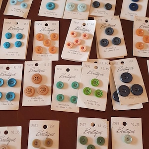 May include: A selection of vintage Boutique brand buttons in diverse colours, such as blue, green, orange, and brown. The buttons are presented on small, labelled cards, with sizes from 13mm to 19mm.