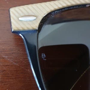 May include: Close-up of sunglasses with black frames and brown lenses. The temples have a light wood grain pattern. A small, silver accent is on the temple. The lenses have a small, etched logo.