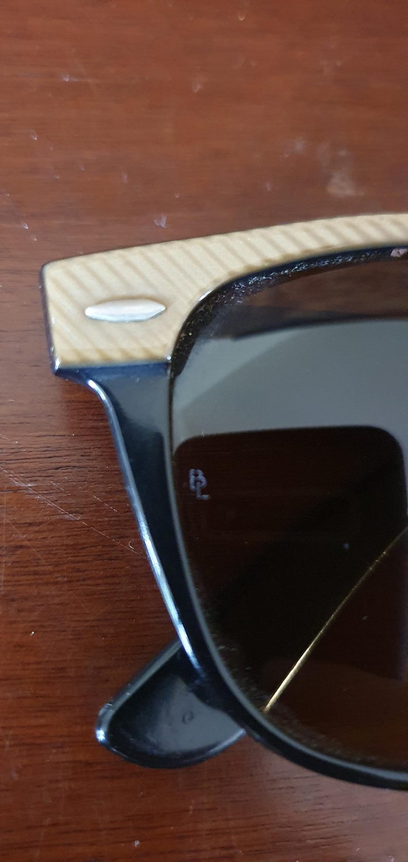 May include: Close-up of sunglasses with black frames and brown lenses. The temples have a light wood grain pattern. A small, silver accent is on the temple. The lenses have a small, etched logo.