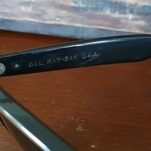 May include: Close-up of black Ray-Ban sunglasses with the text "B&L RAY-BAN U.S.A." printed on the frame. The sunglasses are resting on a wooden surface. The background includes a wooden box with faded text.