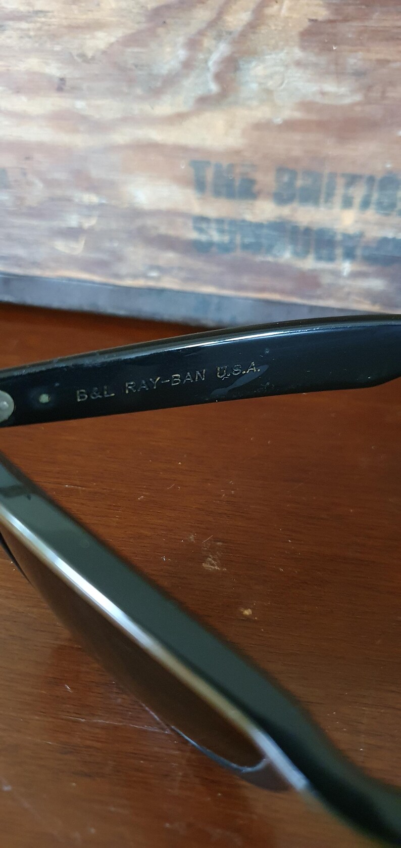 May include: Close-up of black Ray-Ban sunglasses with the text "B&L RAY-BAN U.S.A." printed on the frame. The sunglasses are resting on a wooden surface. The background includes a wooden box with faded text.