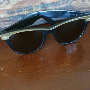 May include: Black cat-eye sunglasses with gold-colored accents on the frames. The sunglasses have dark lenses and are resting on a wooden surface. The text "Ray-Ban USA" is visible on the left arm.