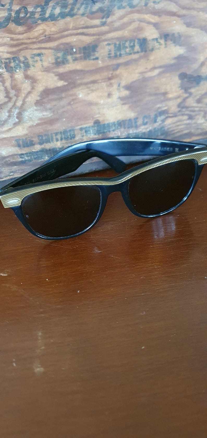 May include: Black cat-eye sunglasses with gold-colored accents on the frames. The sunglasses have dark lenses and are resting on a wooden surface. The text "Ray-Ban USA" is visible on the left arm.