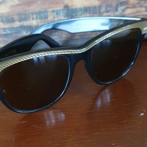 May include: Black cat-eye sunglasses with gold-colored trim. The sunglasses have dark lenses and are set against a wooden background. The frames are a classic style, suitable for various face shapes.