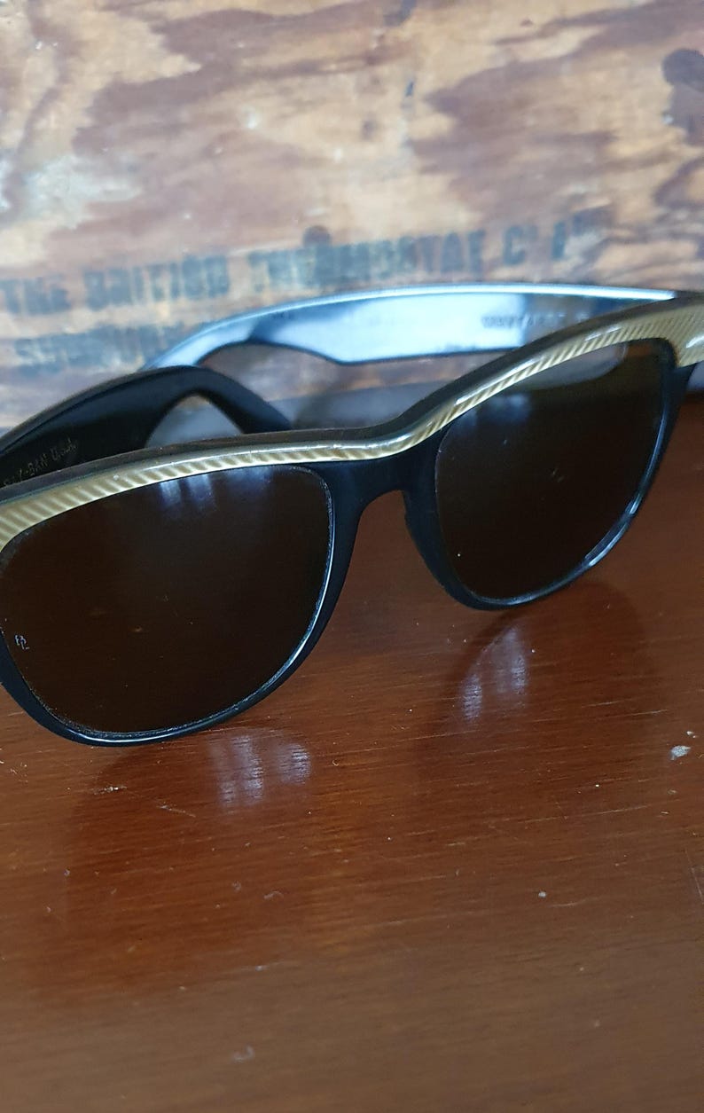 May include: Black cat-eye sunglasses with gold-colored trim. The sunglasses have dark lenses and are set against a wooden background. The frames are a classic style, suitable for various face shapes.