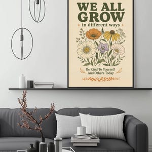 a poster with the text "WE ALL GROW in different ways" and an illustration of flowers, hanging on a wall above a gray couch with a coffee table in front of it.