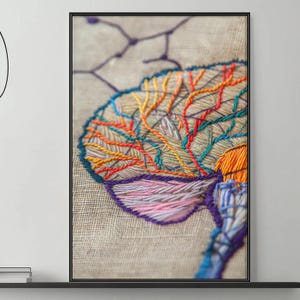 May include: Framed embroidery art featuring a colorful brain design. The artwork showcases intricate stitching in shades of orange, yellow, blue, and pink on a neutral linen background. The piece is framed in a black frame.