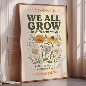 以下が含まれることがあります： WE ALL GROW in different waysおよびBe Kind To Yourself And Others Todayというテキストが書かれた、額装された縦型のウォールアートプリント。オレンジ、黄色、白、紫の花のイラストと、小さな人のシルエットが描かれています。