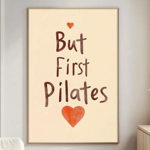 May include: A framed print featuring the text "But First Pilates" in brown, with a small red heart above and a larger red heart below. The background is a light beige colour.