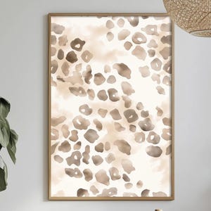 May include: A framed art print featuring a watercolor leopard print design. The artwork showcases a pattern of brown and tan spots against a cream-colored background. The print is encased in a light brown wooden frame, suitable for home decor.