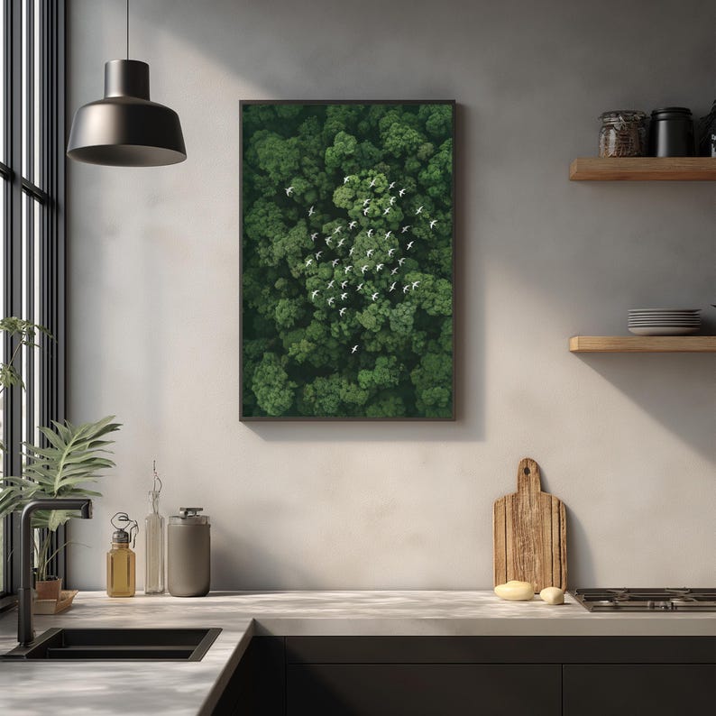 a kitchen counter with various kitchen items, including a cutting board, a sink, and a potted plant. Above the counter, there is a framed picture of a lush green forest with a flock of birds in the sky.