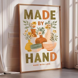 May include: A framed wall art print featuring the text MADE BY HAND and MADE WITH LOVE in orange and olive green. The illustration displays various ceramic vases and bowls in yellow, orange, and green, filled with assorted wildflowers and foliage.