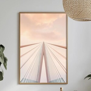 May include: A framed print of a bridge with a central tower and suspension cables. The bridge is painted in shades of pink and white, set against a soft peach-colored sky. The artwork is displayed in a light brown wooden frame.