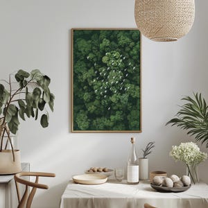 a dining table set with a white tablecloth, a bowl of fruit, a bottle, and a vase with flowers. Above the table, there is a framed picture of a lush green forest with white birds flying in the sky.