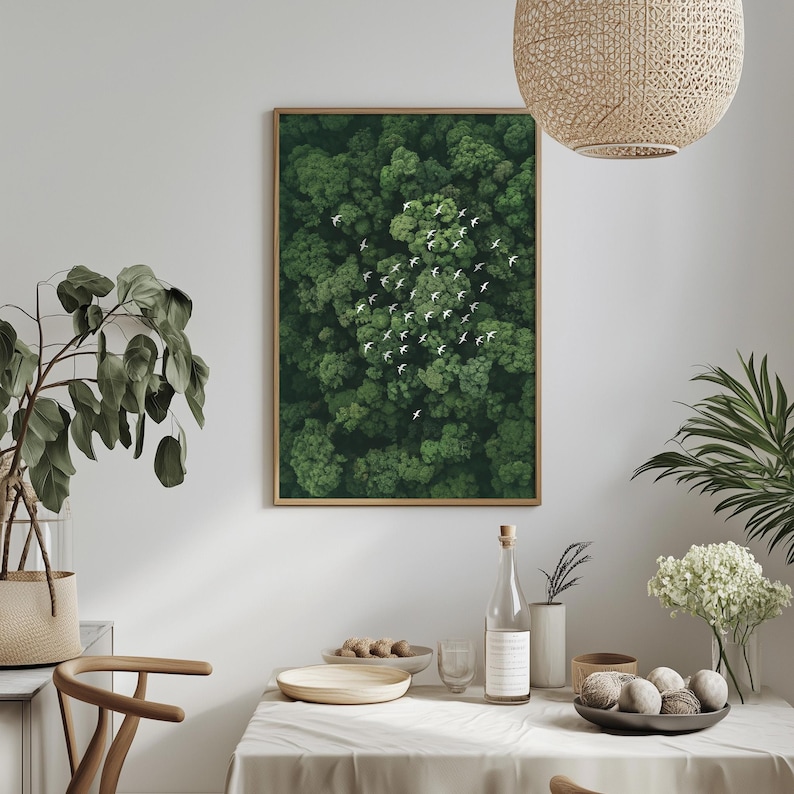 a dining table set with a white tablecloth, a bowl of fruit, a bottle, and a vase with flowers. Above the table, there is a framed picture of a lush green forest with white birds flying in the sky.