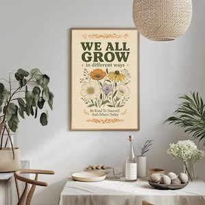 a framed poster with the text "WE ALL GROW in different ways" and illustrations of flowers and leaves. The poster is hung on a wall above a dining table with various items on it, including a bottle, a bowl, and a vase with flowers.