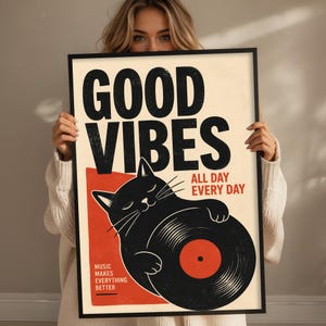 May include: A rectangular framed poster with a cream background featuring a black cat holding a vinyl record. The text reads GOOD VIBES, ALL DAY EVERY DAY, and MUSIC MAKES EVERYTHING BETTER. The design includes a red rectangular accent behind the cat.