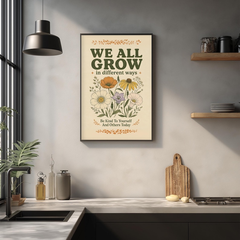 a framed poster with the text "WE ALL GROW in different ways" and an illustration of flowers, hanging on a wall above a kitchen counter with various kitchen items such as a cutting board, a soap dispenser, and a plant.