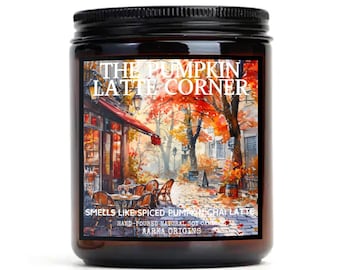 May include: A brown glass jar candle with a black lid. The label features a watercolor illustration of a street scene with a cafe and autumn leaves. The text on the label reads "The Pumpkin Latte Corner" and "Smells like spiced pumpkin chai latte".