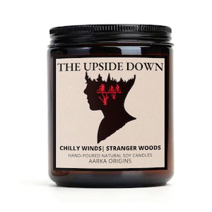 May include: A brown glass jar candle with a black lid. The label is beige with red text that reads "THE UPSIDE DOWN", "CHILLY WINDS | STRANGER WOODS", "HAND-POURED NATURAL SOY CANDLES", and "AARKA ORIGINS". The label also features a black silhouette of a person's head with a forest scene inside.