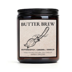 May include: A brown glass candle jar with a black lid. The label is brown with black text that reads "BUTTER BREW" and a black illustration of a broom. The text below the illustration reads "BUTTERSCOTCH | CARAMEL | VANILLA" and "HAND-POURED NATURAL SOY CANDLES AARKA ORIGINS".