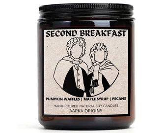 May include: A brown glass candle jar with a black lid. The label features a black and white illustration of two people with the text "SECOND BREAKFAST" above them. The label also includes the text "PUMPKIN WAFFLES | MAPLE SYRUP | PECANS", "HAND-POURED NATURAL SOY CANDLES", and "AARKA ORIGINS".