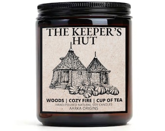 Può includere: Una candela in vetro marrone in un barattolo con un coperchio nero. L'etichetta presenta un'illustrazione in bianco e nero di due piccole case con tetti a punta. Il testo sull'etichetta recita "The Keeper's Hut", "Woods | Cozy Fire | Cup of Tea", "Hand-poured Natural Soy Candles" e "Aarka Origins".