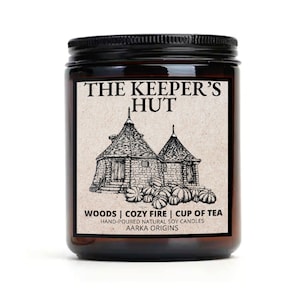 May include: A brown glass jar candle with a black lid. The label features a black and white illustration of two small houses with pointed roofs. The text on the label reads "The Keeper's Hut", "Woods | Cozy Fire | Cup of Tea", "Hand-poured Natural Soy Candles", and "Aarka Origins".