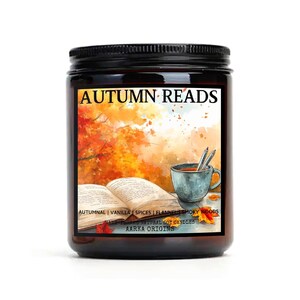 May include: A brown glass candle jar with a black lid. The label features a watercolor illustration of a book, a cup of coffee, and pencils, with a fall-themed background. The text on the label reads "AUTUMN READS", "AUTUMNAL | VANILLA | SPICES | FLANNEL SMOKY WOODS", "HAND POURED NATURAL SOY CANDLES", and "AARKA ORIGINS".